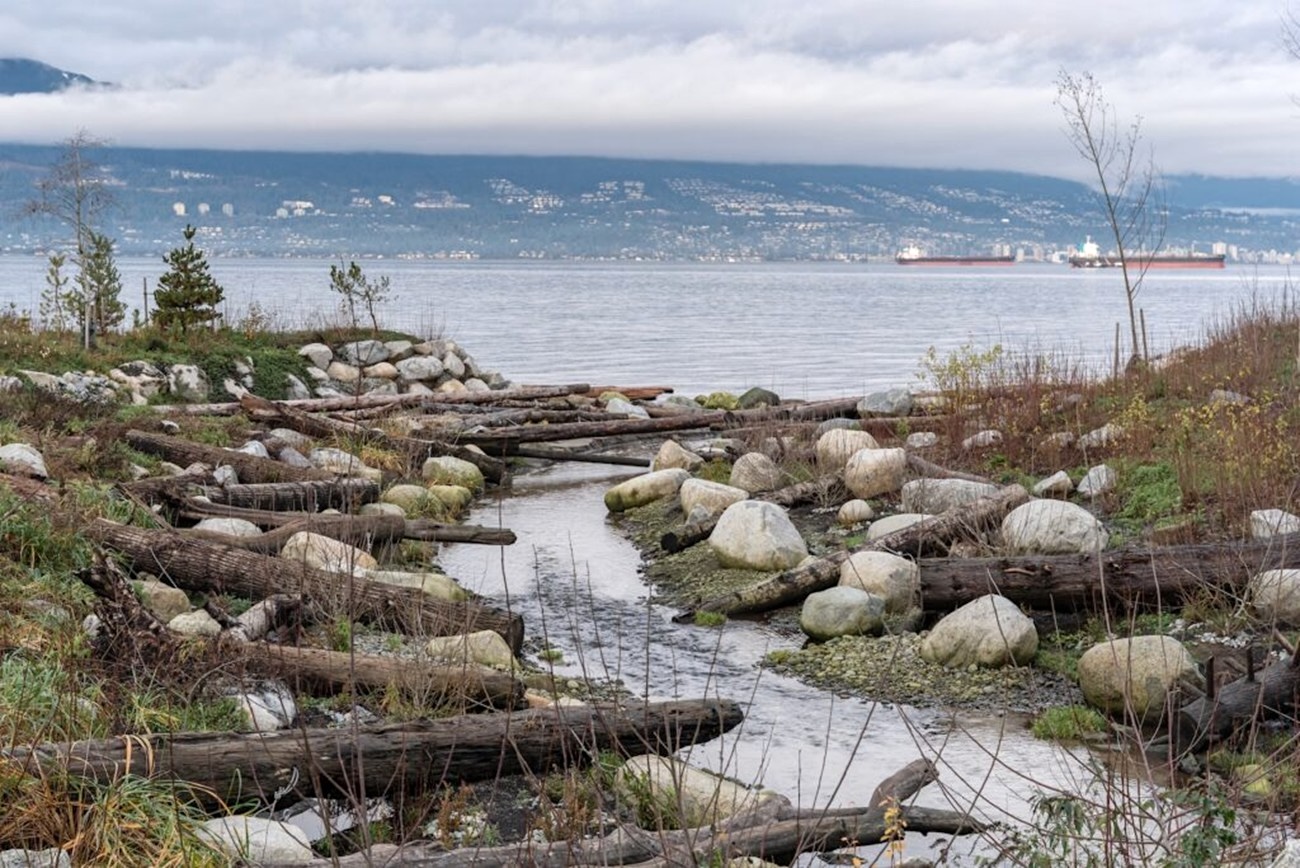 A boulder-lined creek flows into Burrard Inlet, with West Vancouver visible in the distance.
