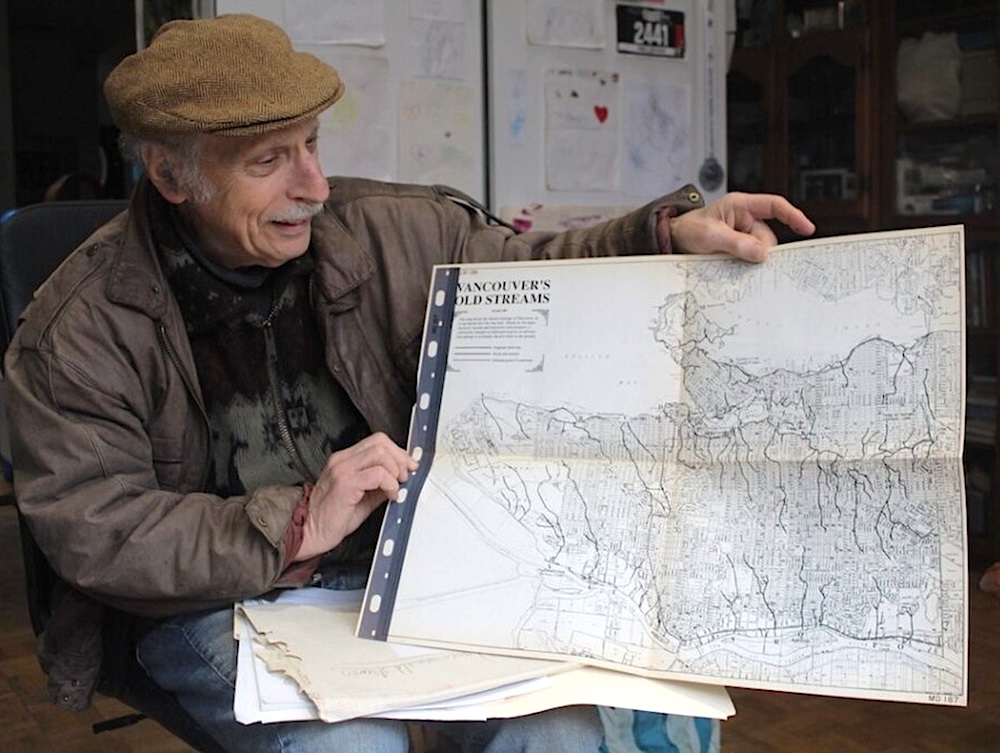 A man with grey hair and moustache wearing a tweed flat cap holds up a map titled &lsquo;Vancouver&rsquo;s Old Streams.&rsquo;