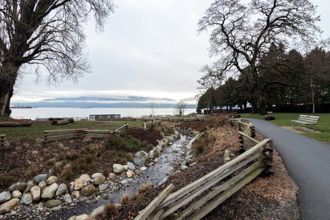 A small boulder-lined creek flows through an urban park toward Burrard Inlet visible in the distance.
