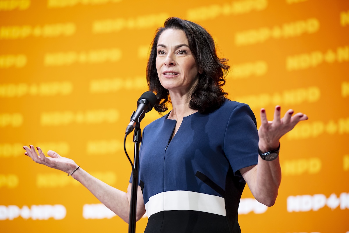 A woman with light skin and shoulder-length brown hair stands behind a microphone.
