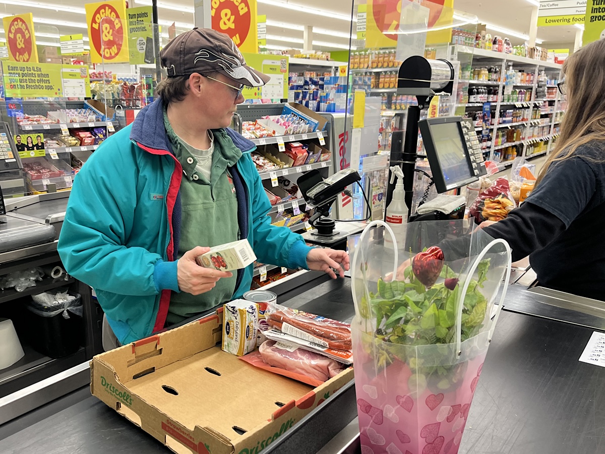 A man with light skin tone wearing a ball cap stands at the checkout of a grocery store.
