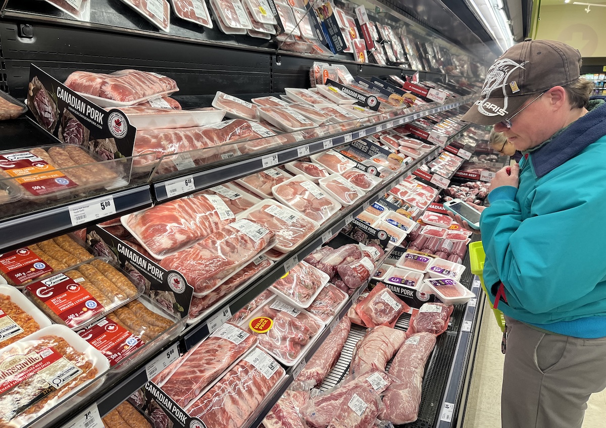 A man with light skin tone wearing a ball cap Looks over the meat section in a grocery store.