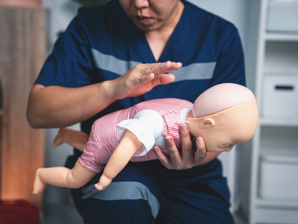 A pediatric first aid trainer with dark hair and medium skin tone demonstrates using back blows on a baby mannequin.