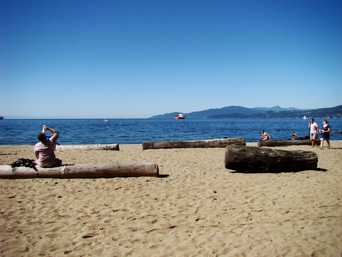 A sandy beach with logs and a few people on a sunny day. A cargo ship and some sailboats are visible in the distance.
