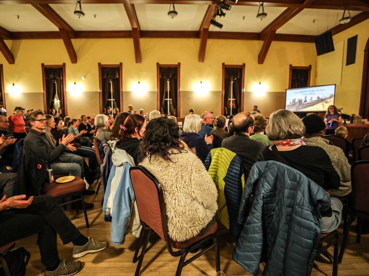 A large number of seated people take in a community meeting at a local hall.