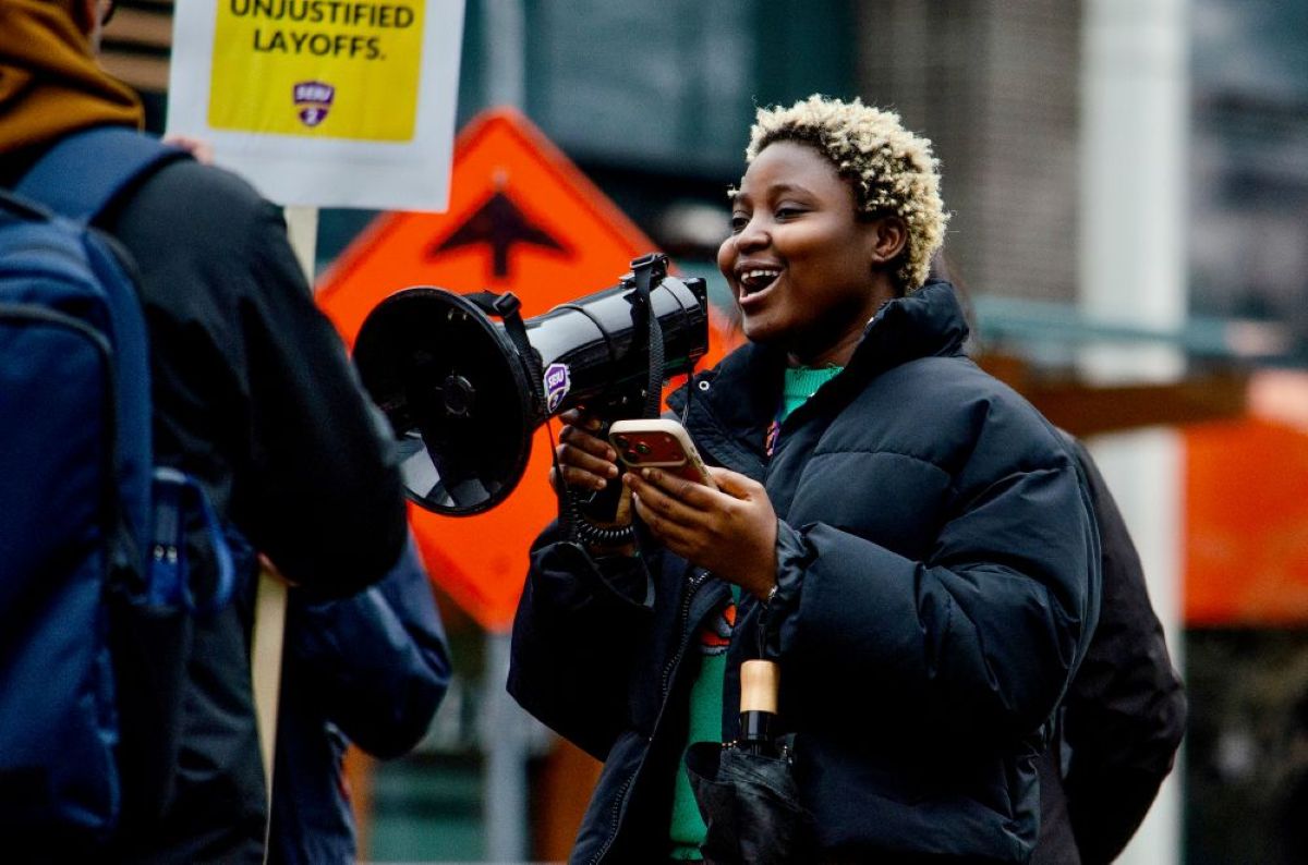 A young woman with dark skin and bleach-tipped short hair stands with a megaphone at a rally outdoors. She wears a puffy black coat and holds an iPhone in one hand.