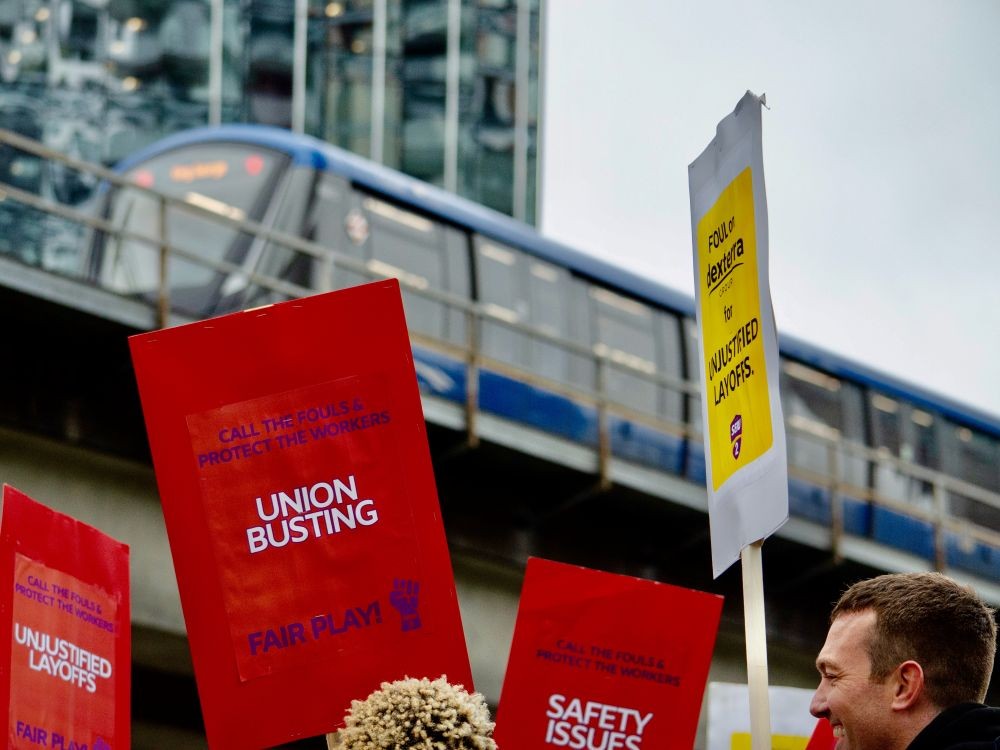 A group of people carry red signs that read &ldquo;call the fouls and protect the workers &mdash; union busting&rdquo; as a SkyTrain passes by overhead.