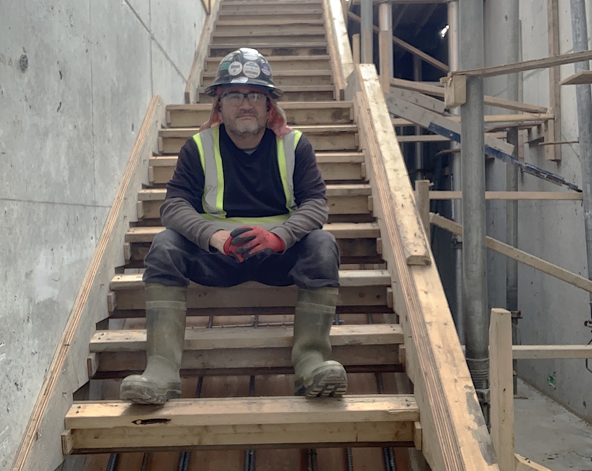 Francisco Barahona sits on stairs at a work site. He is wearing a hard hat and other construction equipment.