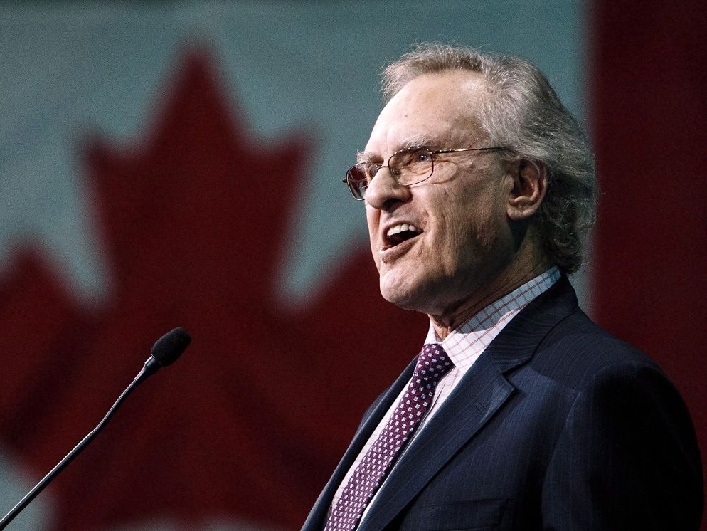 Seen in profile, a man in late middle age with light skin and grey hair, wearing a suit and tie, speaks into a microphone. A large Canadian flag can be seen in the background.