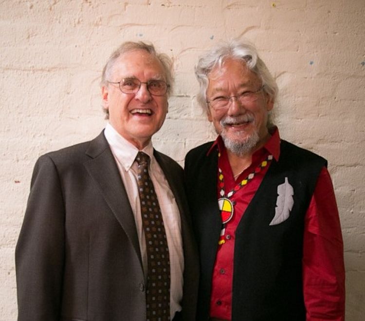Two older men stand side by side smiling. On the left, Stephen Lewis has light skin and thinning grey hair and wears a dark brown jacket and tie. On the right, David Suzuki has medium-light skin tone and silver hair and goatee, and wears a black vest over a red shirt.