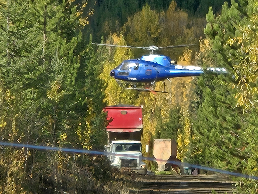 A helicopter is hovering near the ground. Below it are large trucks and other equipment.