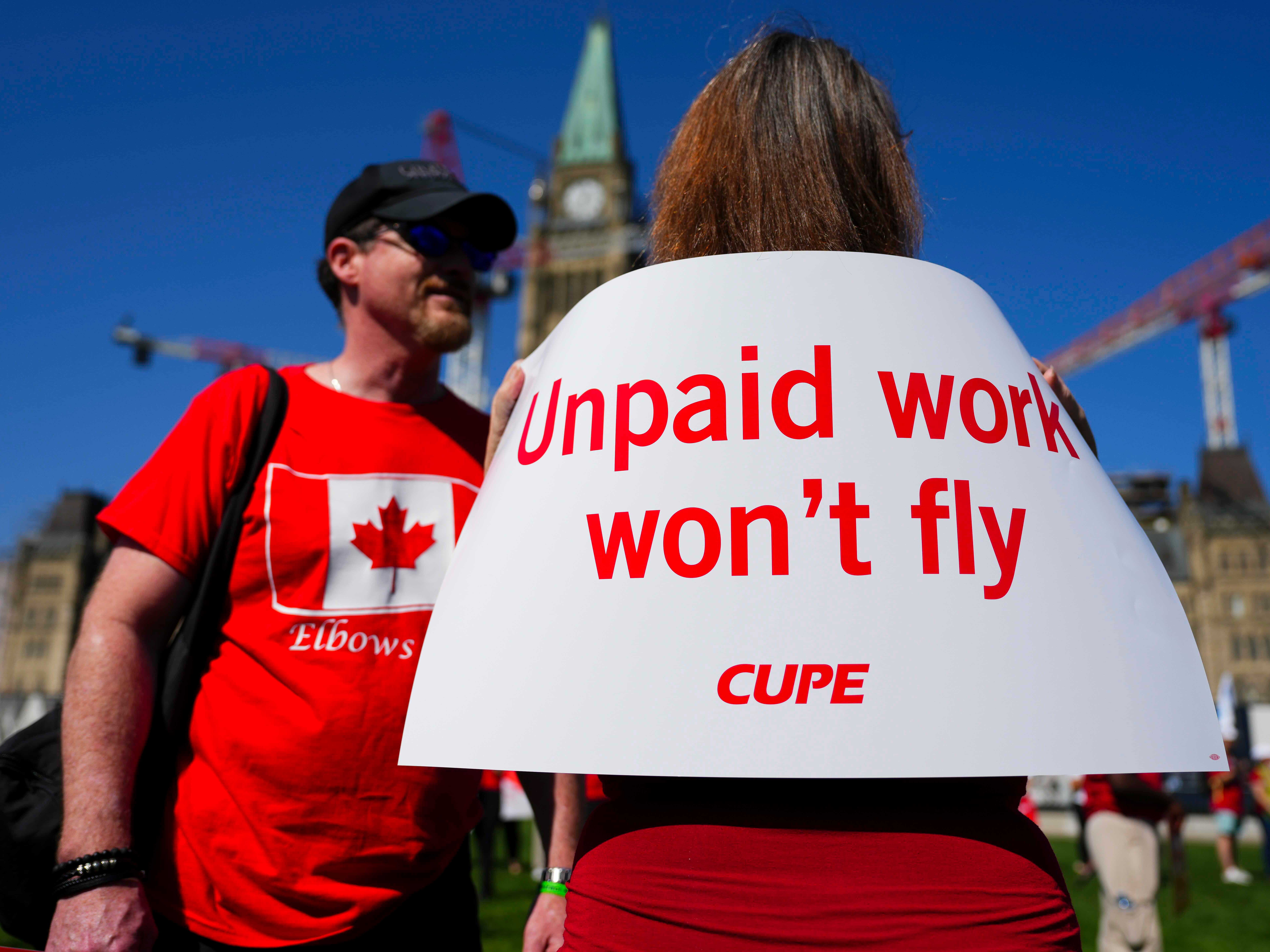 A woman dressed in a red T-shirt stands with her back to the camera, displaying a sign that reads &ldquo;unpaid work won&rsquo;t fly.&rdquo; Beside her is another person dressed in a red T-shirt decorated with a Canadian flag. The top of the Peace Tower at the Canadian Parliament can be seen in the background.