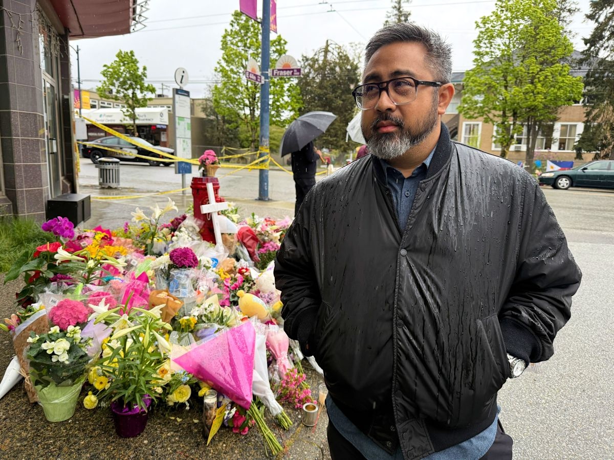 RJ Aquino is a middle-aged man with black-rimmed glasses and dark hair, wearing a black leather jacket on a street corner. Behind him is a collection of flowers left as a memorial.