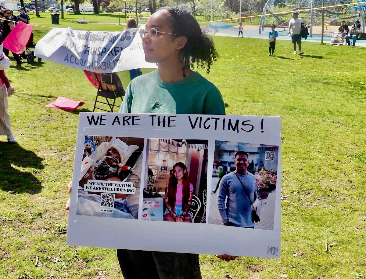 Vanessa Hill has curly dark hair tied back. She is wearing gold-rimmed glasses and a green shirt, and standing in a grassy area holding a homemade poster that reads, &lsquo;We are the victims!&rsquo;