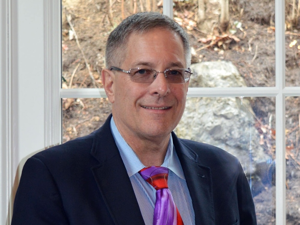 A light-skinned man with glasses wearing a suit and a purple and red tie.