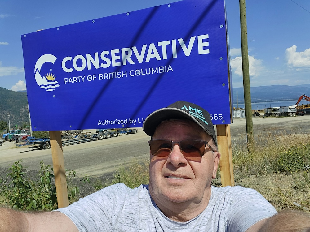 A middle-aged man with light skin tone is wearing a ball cap and taking a selfie in front of a Conservative Party of BC sign. 