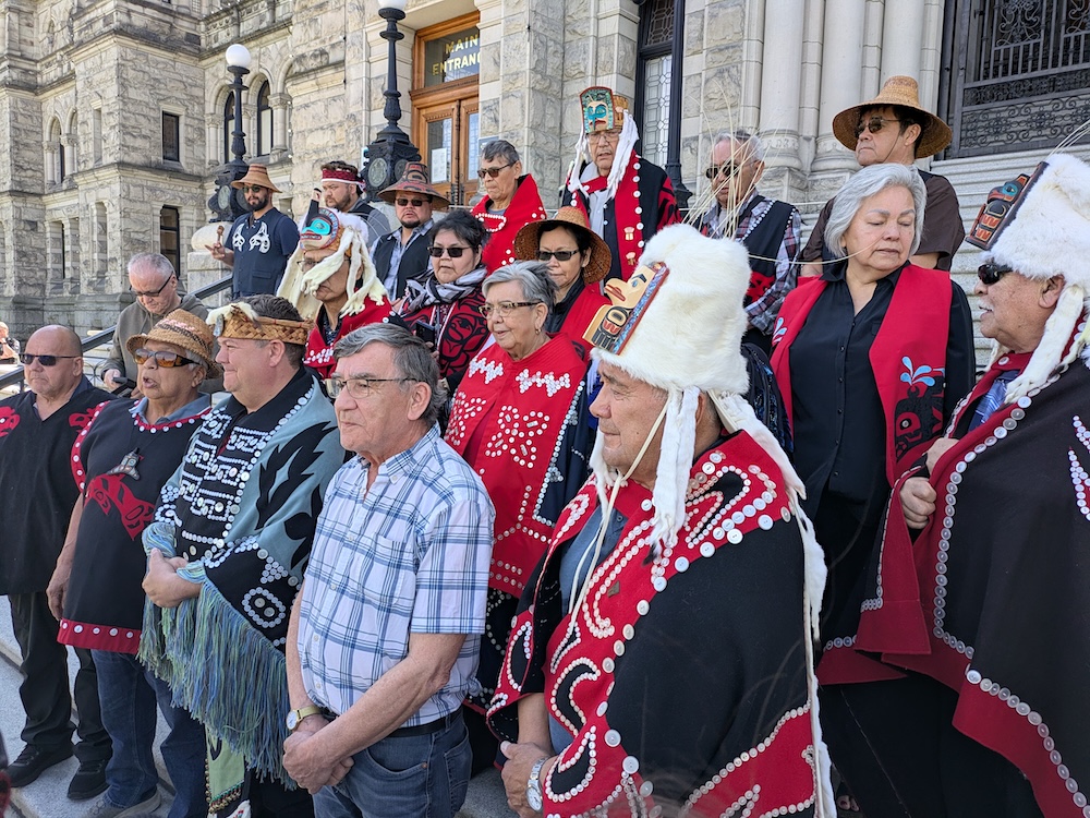 About two dozen people, most in traditional regalia, stand on the stone steps of the legislature.