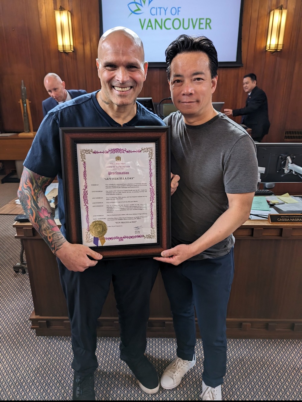 Guy Felicella, a bald man with light skin and arm tattoos hold a plaque while standing next to Mayor Ken Sim, a man with medium-light skin and dark hair wearing a T-shirt, casual pants and sneakers.