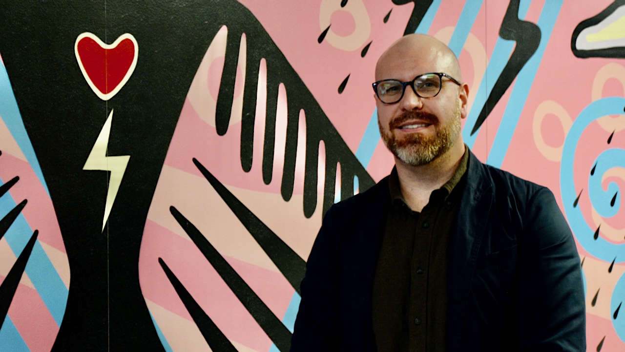 A man with light skin, wearing glasses and a beard, sits in front of a brightly coloured pink and blue mural.