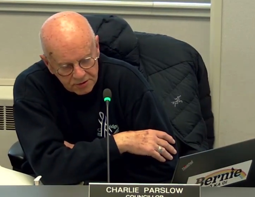 A light-skinned man with no hair and glasses and wearing a sweater speaks into a thin microphone. His laptop has a large &lsquo;Bernie&rsquo; sticker and a rainbow.