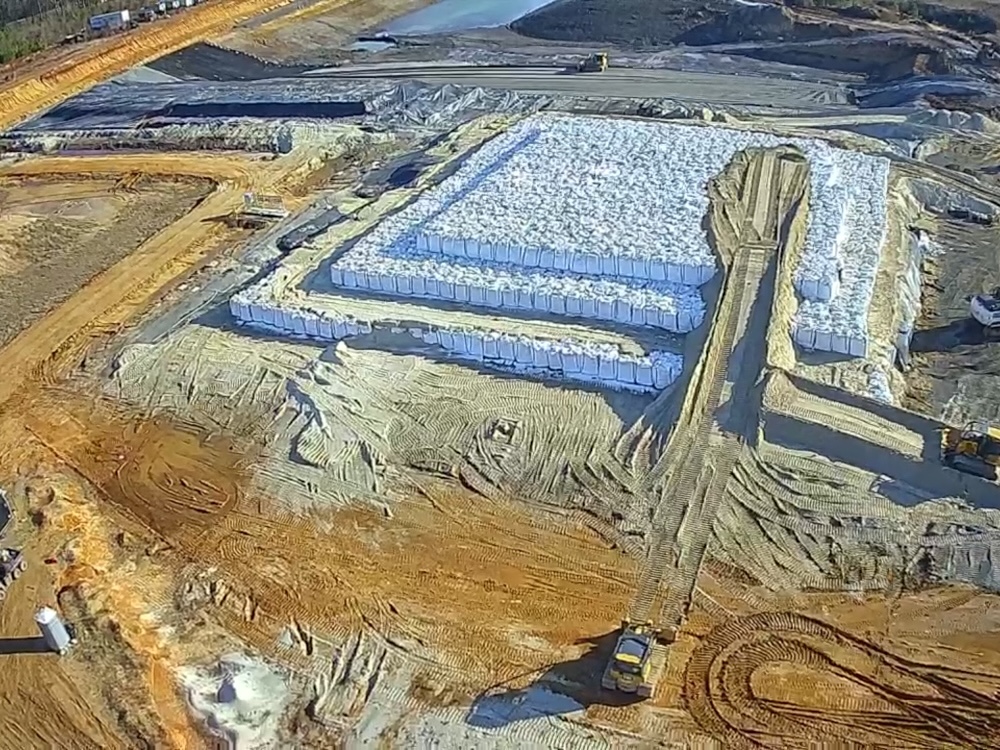 An aerial image shows white bags or sacks arranged in a tiered, square formation. There are three tiers, and a bulldozer has created a dirt road to allow for a fourth tier to be added. 
