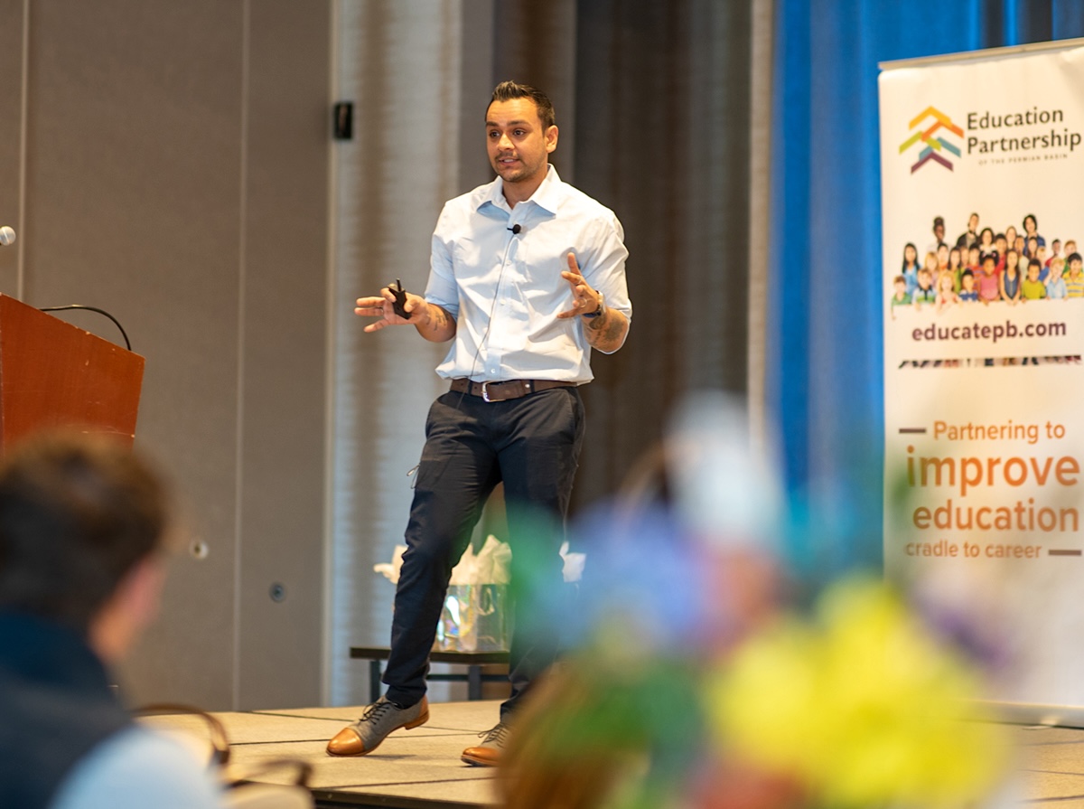 A man with medium-light skin tone and short dark hair wears trousers and a collared shirt. He is addressing an audience at an education event.