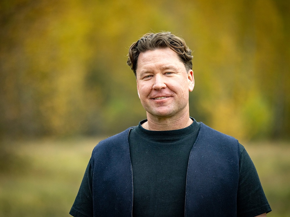 A man with short hair and wearing dark clothing stands in a forest and smiles.