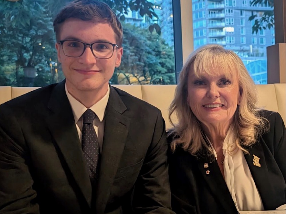 A young man with short brown hair and glasses sits beside an older woman with long wavy blond hair and bangs.