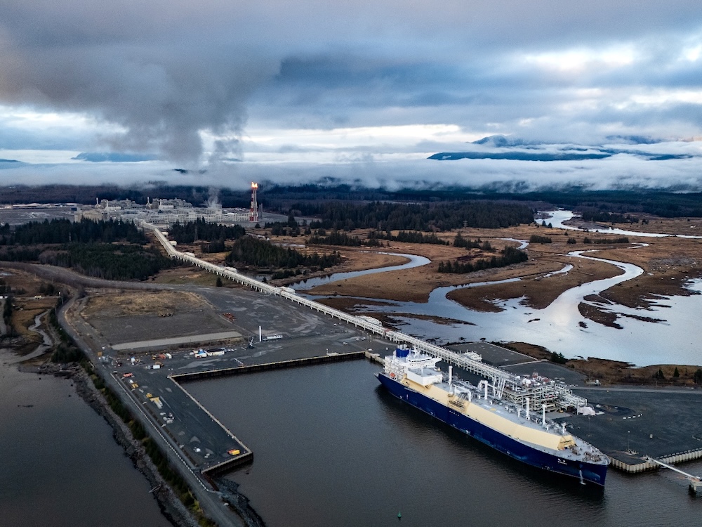 A tanker ship sits at a loading terminal with an estuary and mountain range in the background.