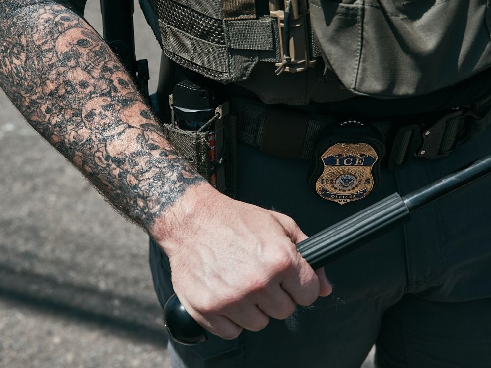 A man&rsquo;s hand grips a police baton. Numerous overlapping skull tattoos can be seen on his forearm, and he wears an olive green uniform with an &lsquo;ICE officer&rsquo; badge and a can of pepper spray attached to his belt.