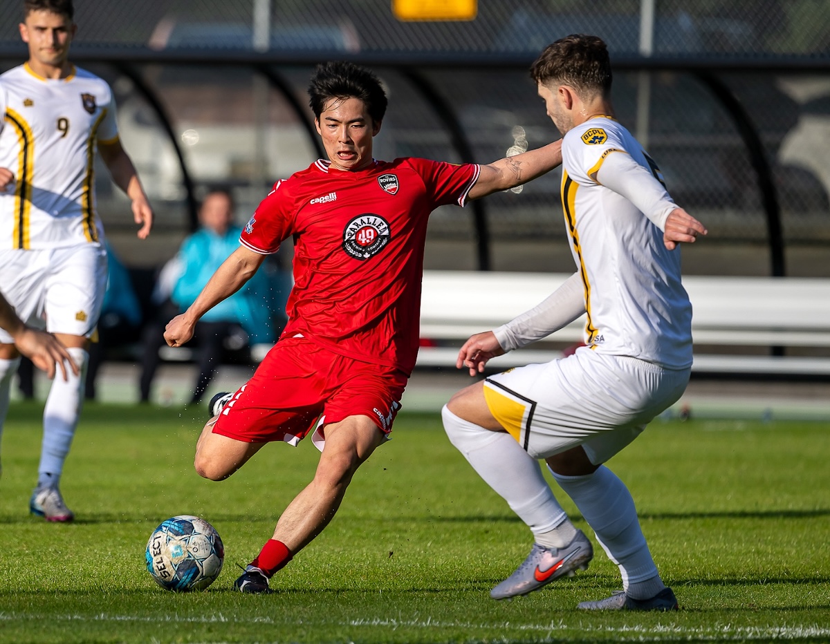 A soccer player in a red uniform prepares to strike a ball as another player, in a white uniform, plays defence.