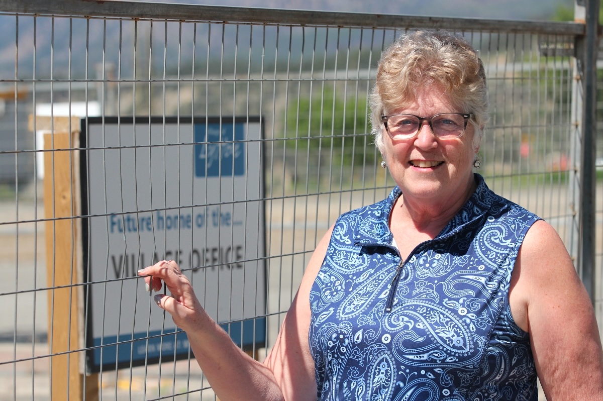 A woman with light skin tone and short blond-grey hair wears a floral shirt and stands with her hand on a construction fence. Behind her, a sign reads, &lsquo;Future home of the... Village Office.&rsquo;