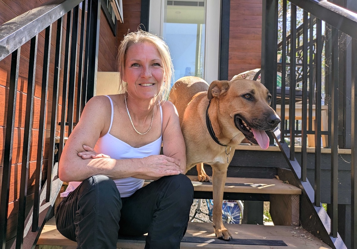 A woman with light skin tone and blond hair pulled back into a ponytail sits on wooden steps with her dog.