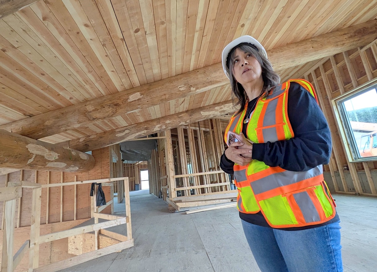 A woman with medium-light skin tone and shoulder-length grey hair wears a high-vis vest and a hard hat and stands in a building under construction.