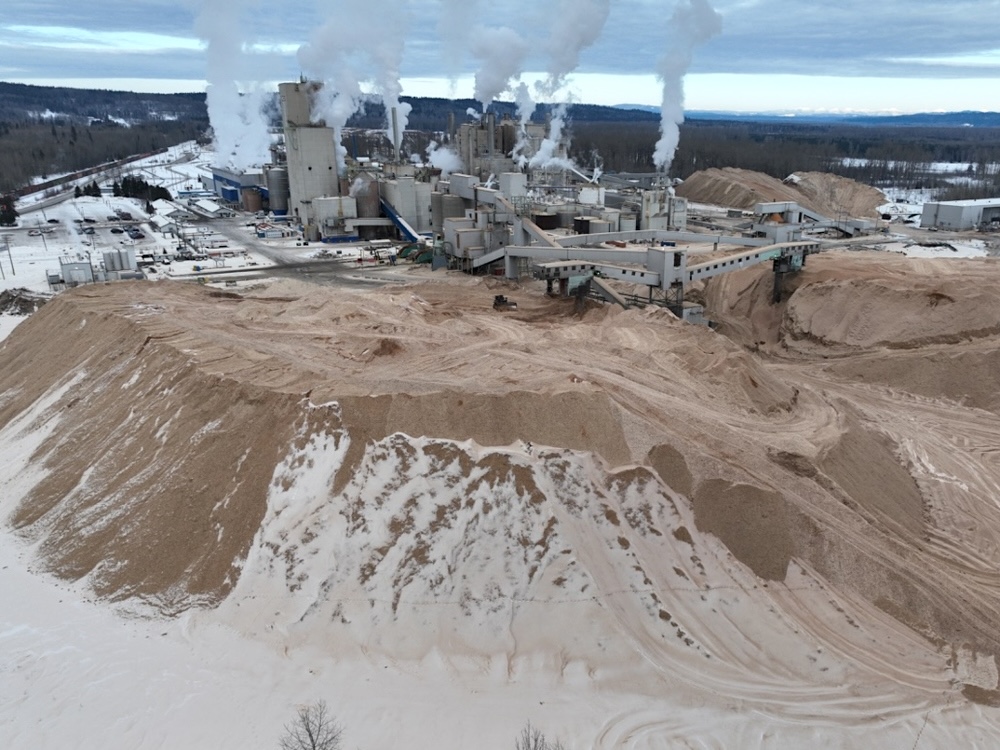 A large plateau-pile of fine-grained wood fibre sits partially covered by snow near a large industrial operation.