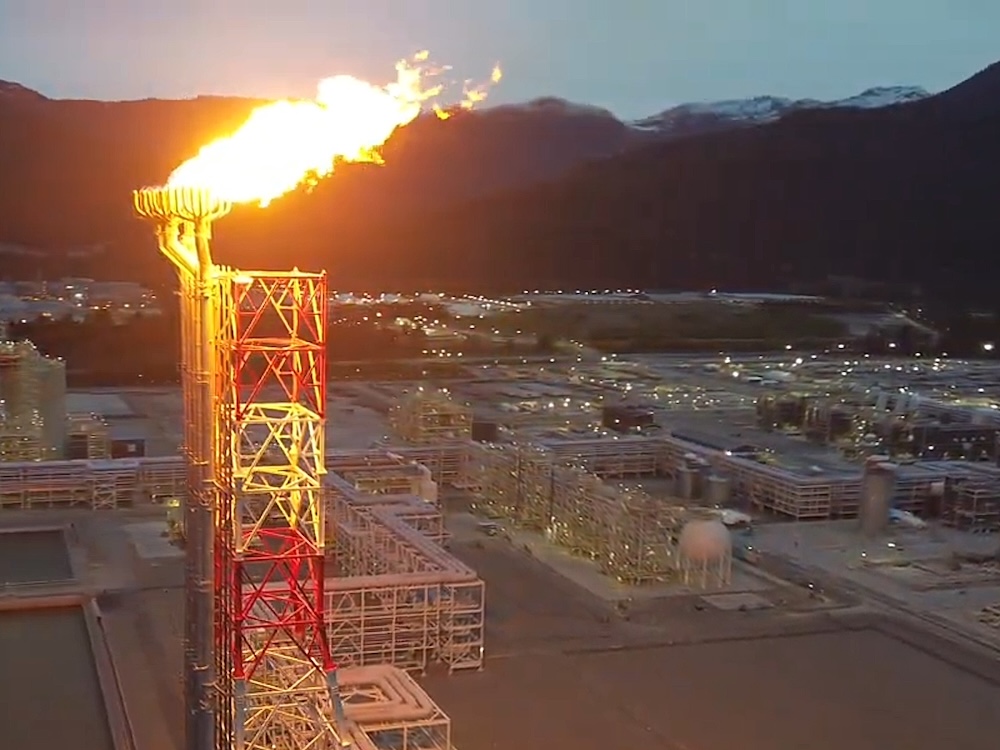 A large flame is emitted from a tower. In the background is a large industrial facility and a mountainous backdrop.