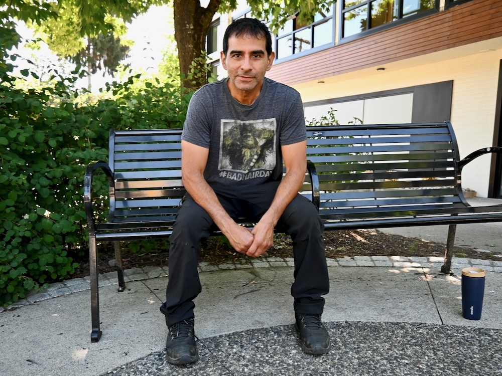 A man with black hair leans forward while sitting on a park bench.