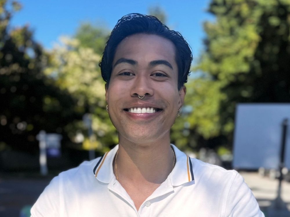 A man with medium-skin, wearing a white polo shirt, smiles for the camera on a sunny day in a place with some deciduous trees.