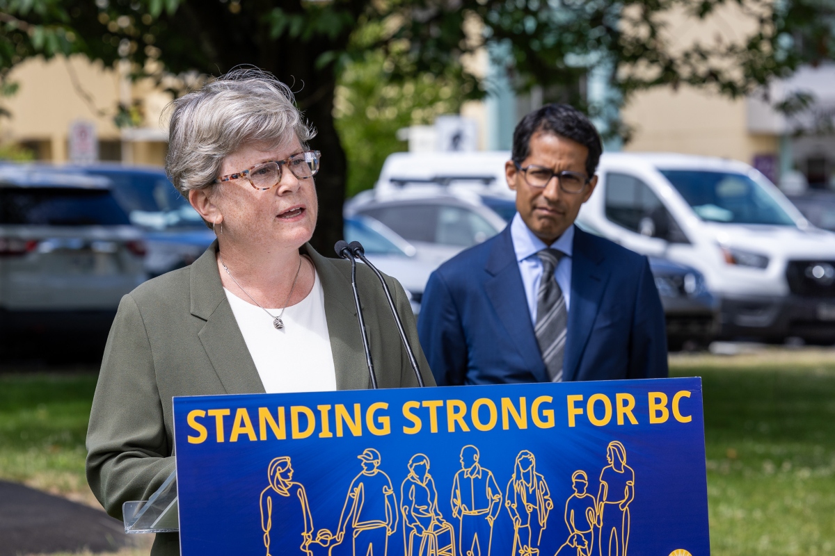 A light-skinned woman with greying hair and glasses stands before a podium bearing the words &ldquo;Standing Strong For BC.&rdquo; A man in a blue suit looks on.