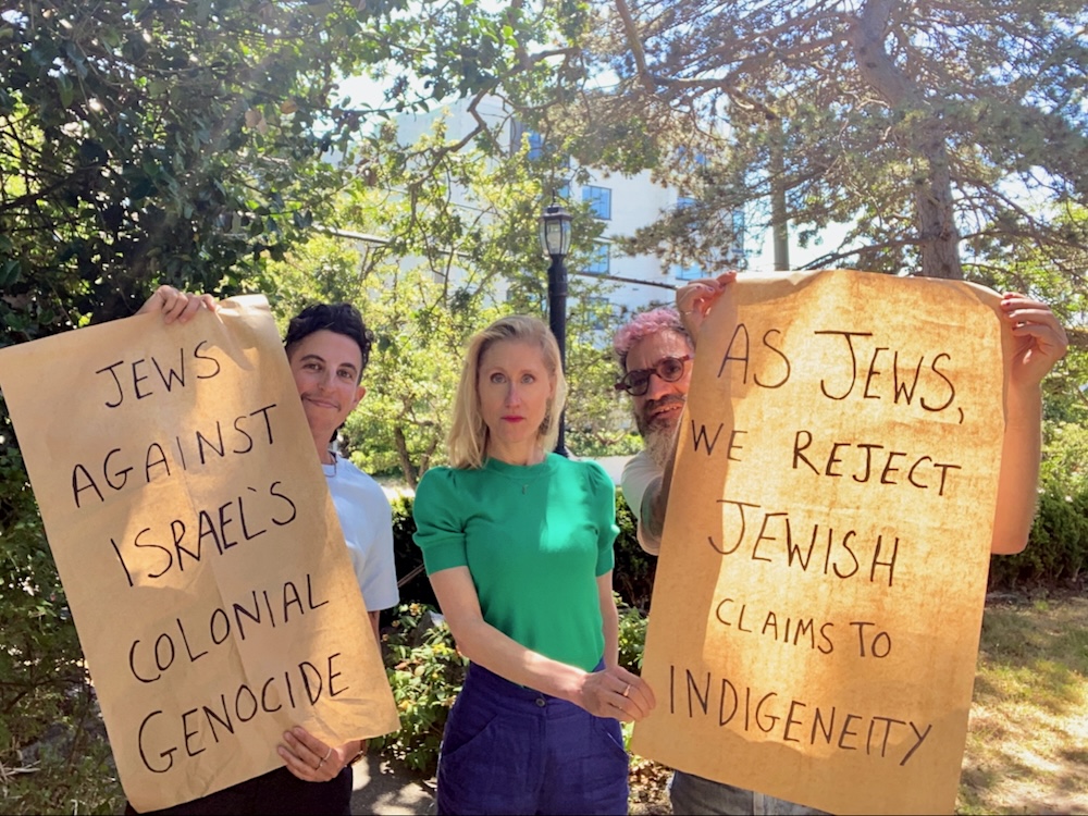 Three people in an outdoor setting hold up posters that read, &lsquo;Jews against Israel&rsquo;s colonial genocide&rdquo; and &ldquo;As Jews, we reject Jewish claims to indigeneity.&rsquo;