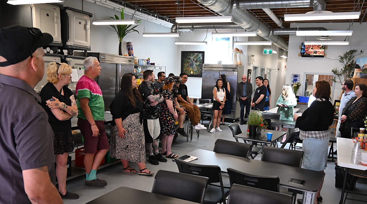 A little over a dozen people stand in a brightly lit space with tables in the foreground and food storage in the background. There are paintings hung around the space, and skylights letting in natural light. 