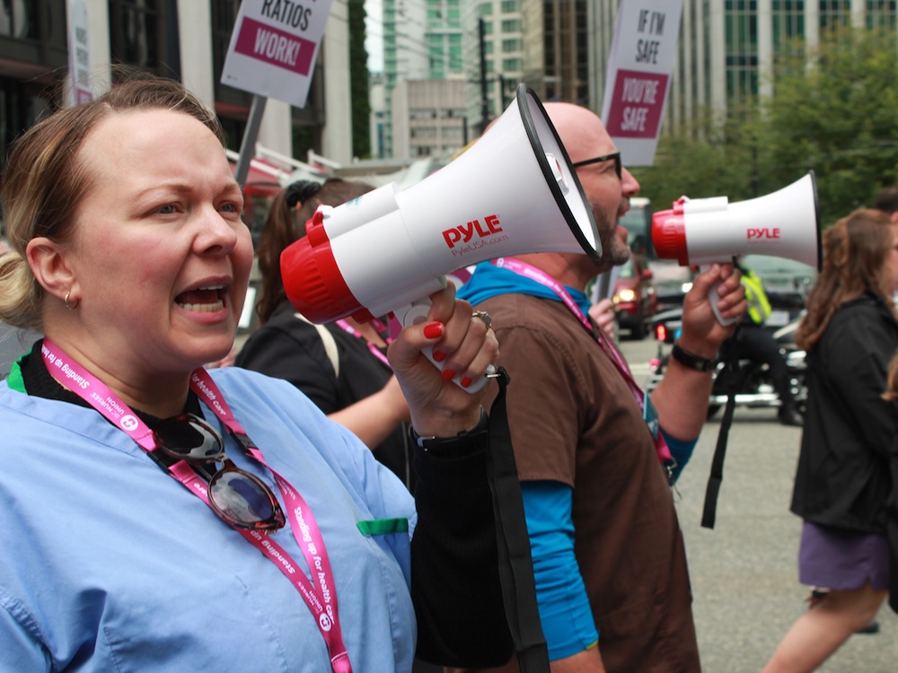 A woman with light skin tone and brown hair in a ponytail wears blue scrubs and pink lanyard. She speaks into a white and red bullhorn at a rally.