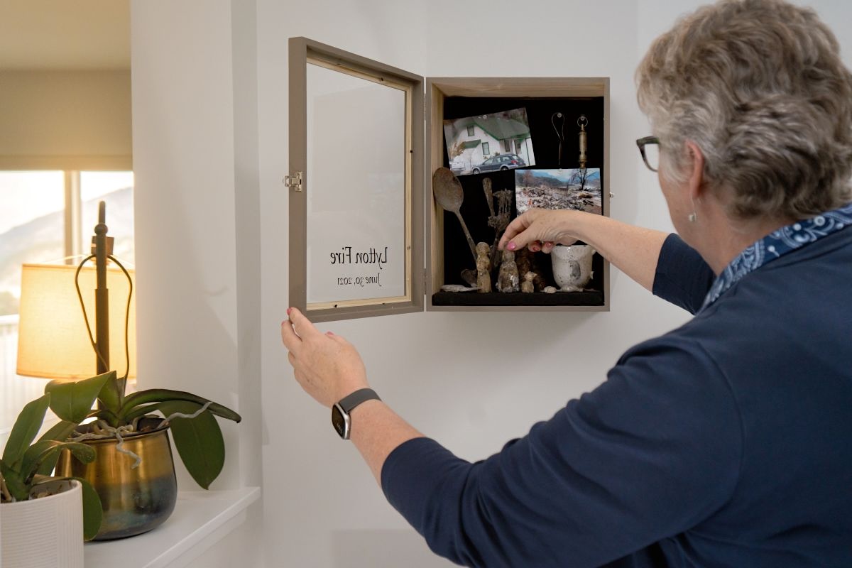 A woman seen from behind, with short grey hair and glasses, opens a small wooden wall-mounted cabinet. The cabinet door, now swung open, is labelled "Lytton Fire, June 30, 2021" in cursive text visible as a mirror image. Inside the cabinet, mounted against a black background, are charred and melted objects, two small photographs &mdash; one showing a house with a car parked outside, another showing a landscape of ash and debris &mdash; and what appears to be a brass fitting or fixture.