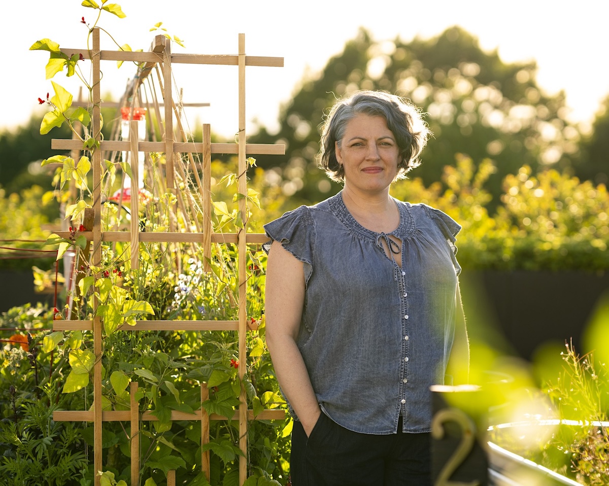 A woman with white skin and short wavy grey-and-dark hair wearing a grey-blue sleeveless button-up top and dark pants stands with hands in pockets beside a wooden trellis wound with climbing plants in a rooftop community garden. Behind her are raised planters, other plots and trees in warm backlit evening light.