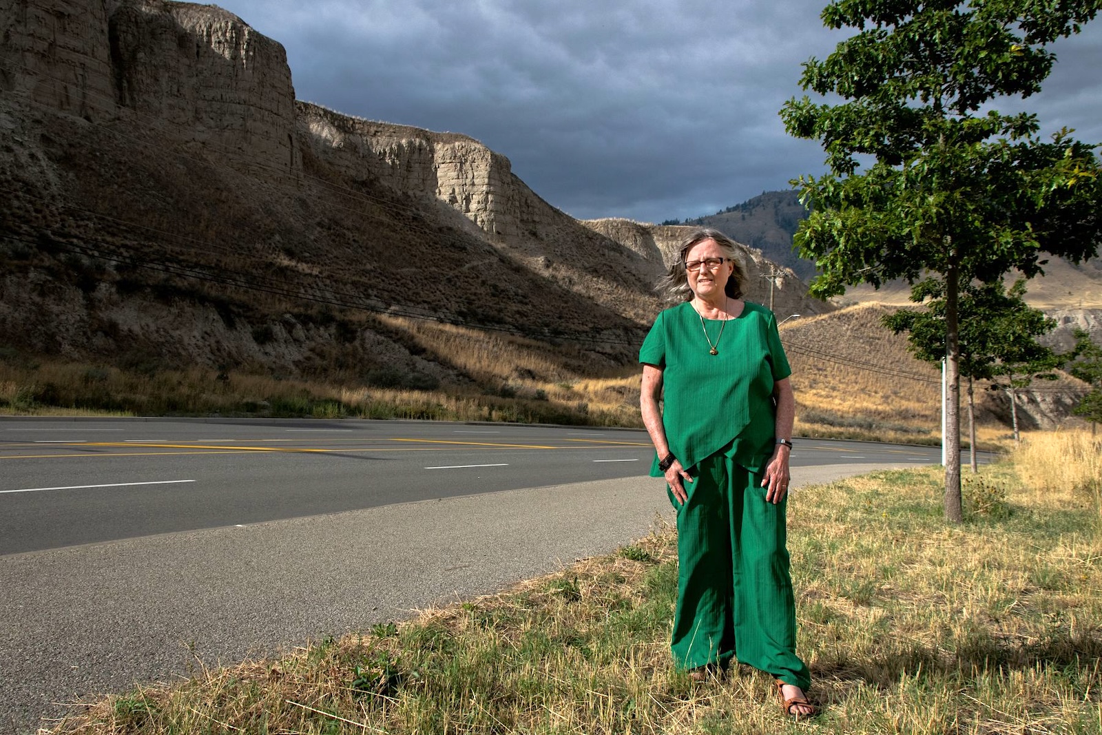 A woman with shoulder-length grey hair and glasses, wearing a green top and matching wide-leg pants, stands on a grassy roadside shoulder. Behind her, tall eroded sandstone cliffs rise under a dark, cloudy sky, with a tree and stretch of empty highway to her right.