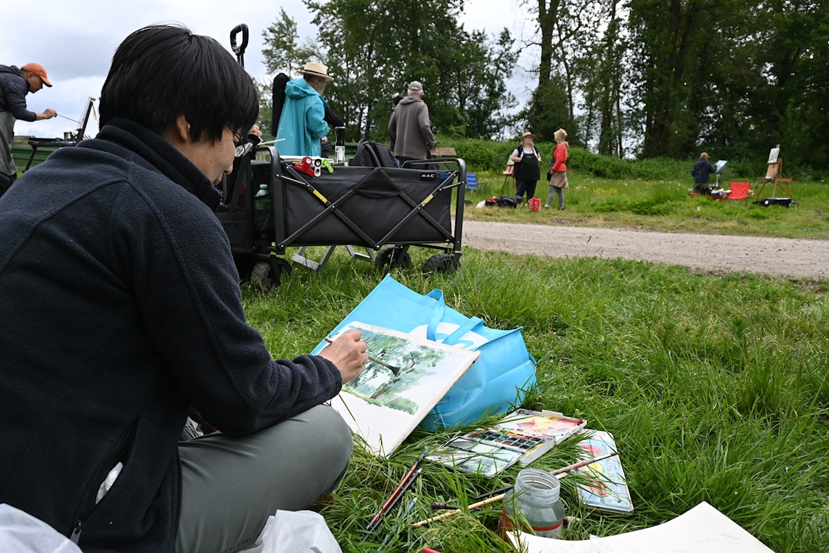 A woman with short dark hair and a black fleece jacket sits in the long grass. Watercolour paints and paintbrushes are gathered in front of her. She is painting trees on a canvas leaning against a blue bag. 