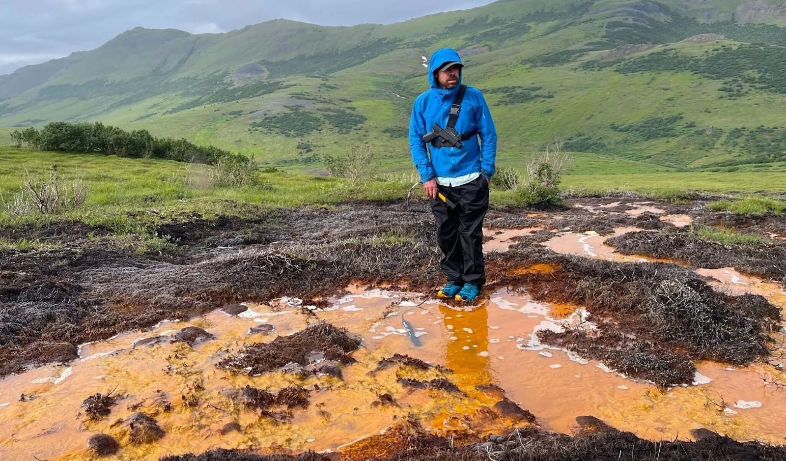 A person in a blue jacket stands beside a muddy brown patch of water, with grassy mountains in the background.