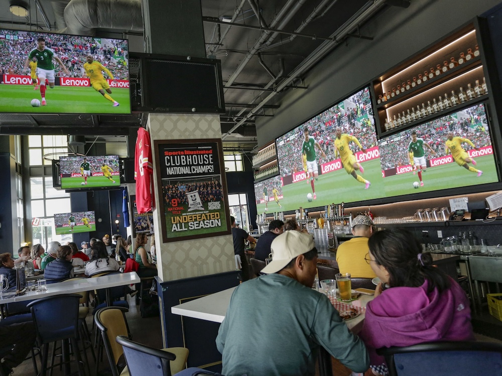People watch soccer on large TV screens in a restaurant with large windows and high ceilings.