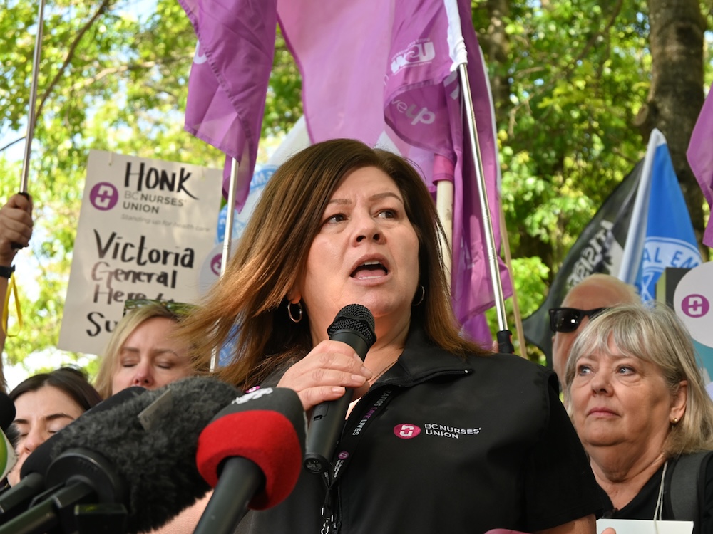 Adriane Gear, a woman with light skin tone and shoulder-length brown hair, speaks into a microphone. She is flanked by picketers and supporters holding signs.