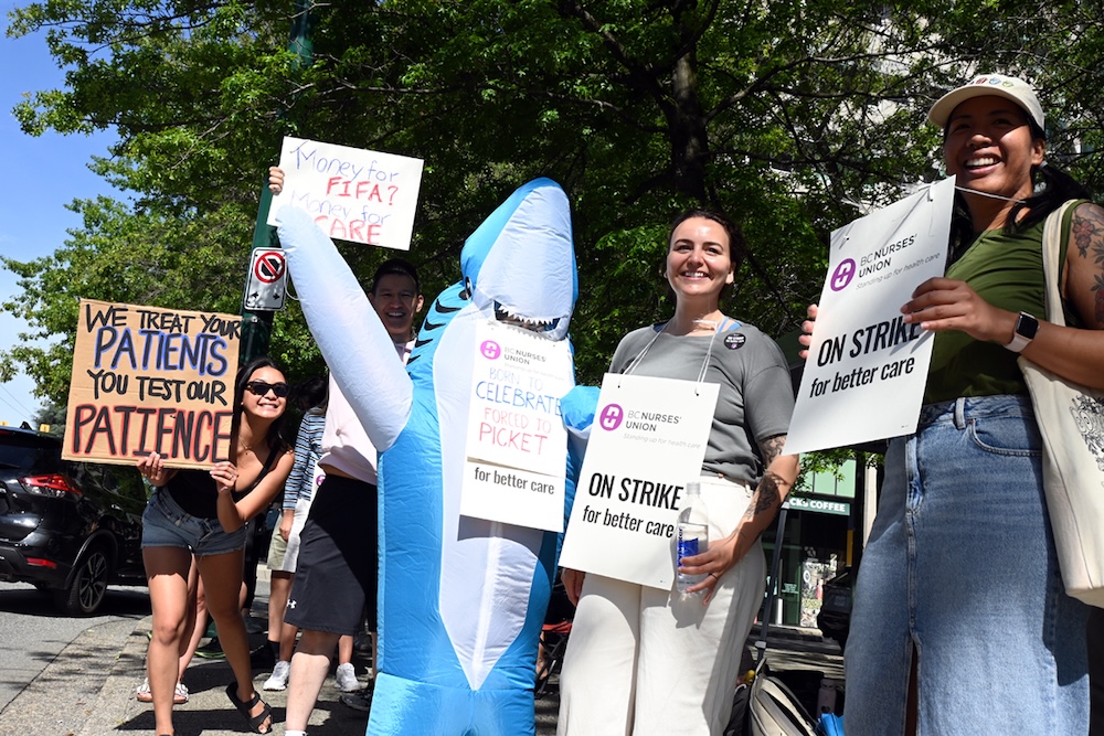 Nurses union picketers hold picket signs. One says, &lsquo;We treat your patients. You test our patience.&rsquo; One wears an inflatable shark costume.